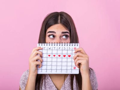 Portrait of a funny young girl in hiding behind a menstrual periods calendar and looking away at copy space isolated over pink background. Female Period calendar